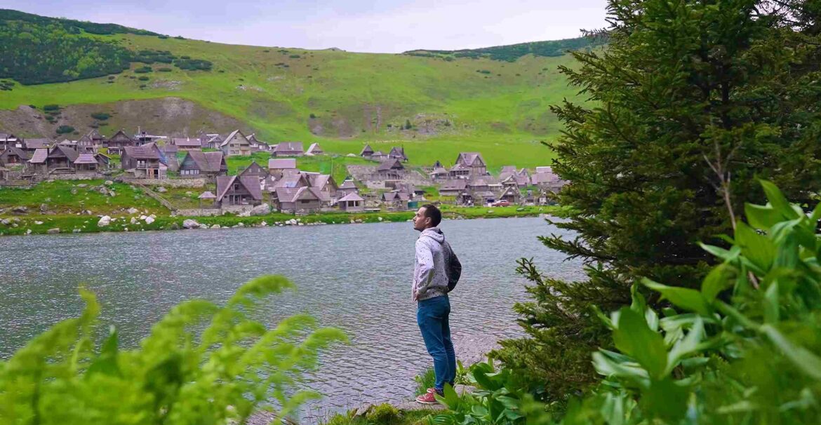 Čovek stoji pored planinskog jezera sa tradicionalnim kućicama i zelenim brdima u pozadini.