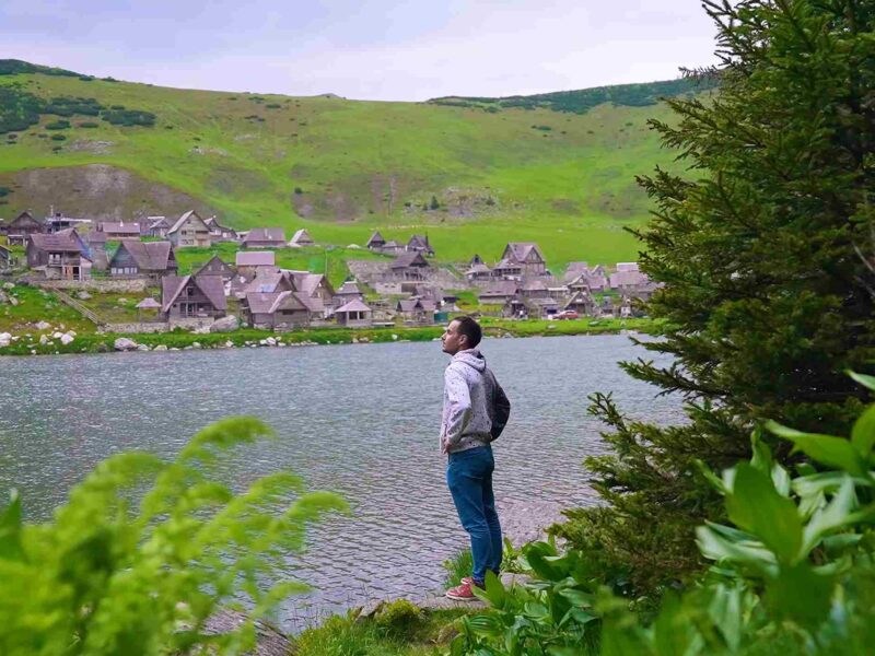 Čovek stoji pored planinskog jezera sa tradicionalnim kućicama i zelenim brdima u pozadini.